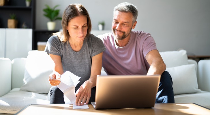 A man and a woman sit on a couch in front of a laptop computer on a coffee table. 