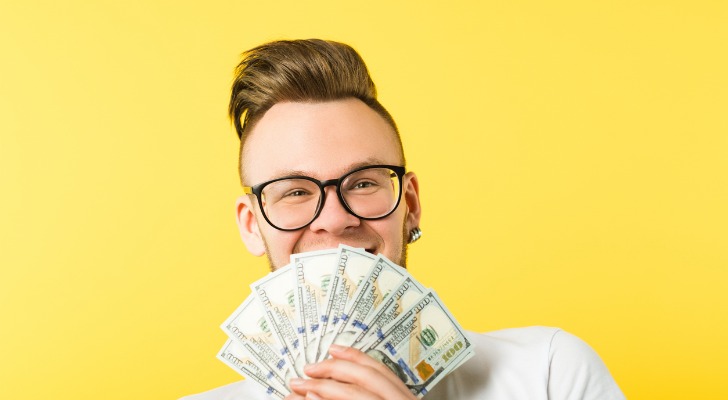 A man in his 20s or 30s smiles while holding $100 bills in front of his face. 